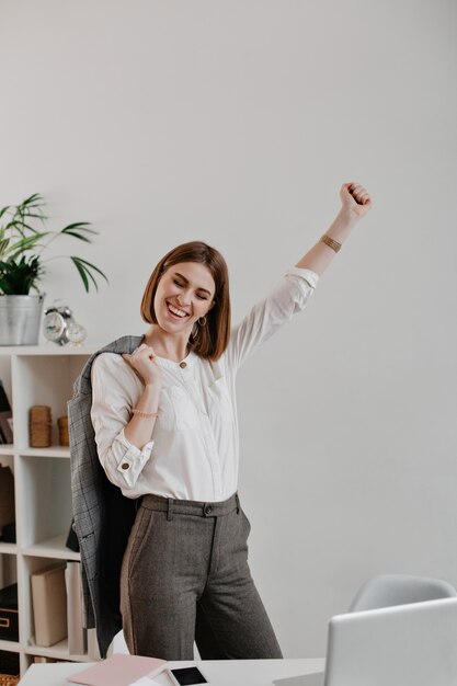 Portrait of young attractive woman in office outfit enjoys business success against bright workplace.