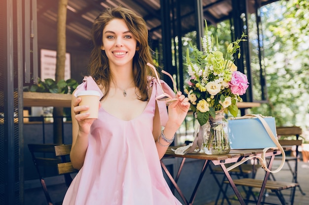 Portrait of young attractive woman sitting in cafe, summer fashion outfit, hipster style, pink cotton dress, sunglasses, smiling, drinking coffee, stylish accessories, trendy apparel, happy mood