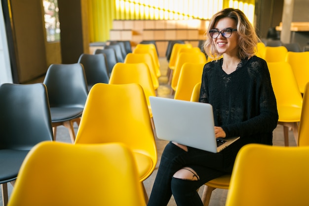 Portrait of young attractive woman sitting in lecture hall, working on laptop, wearing glasses, classroom with many yellow chairs, student learning, education online, freelancer, happy, smiling