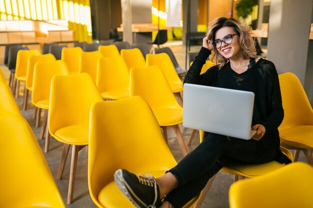Portrait of young attractive woman sitting in lecture hall working on laptop wearing glasses, student learning in classroom with many yellow chairs