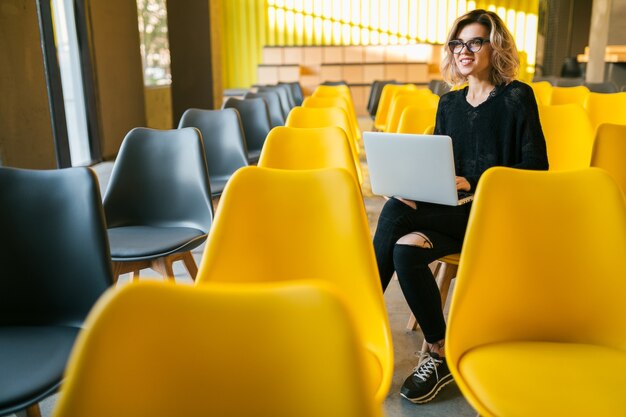 眼鏡をかけているラップトップに取り組んでいる講堂に座っている若い魅力的な女性の肖像画、多くの黄色い椅子のある教室で学ぶ学生