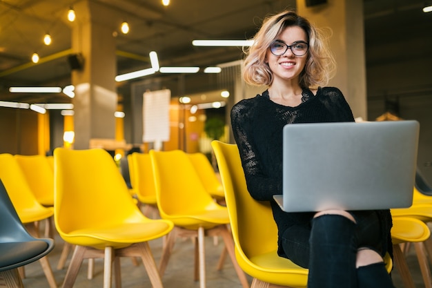 안경을 쓰고 노트북에서 일하는 강당에 앉아있는 젊은 매력적인 여자의 초상화, 많은 노란 의자가있는 교실에서 학습하는 학생