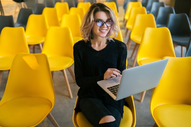 Portrait of young attractive woman sitting in lecture hall working on laptop wearing glasses