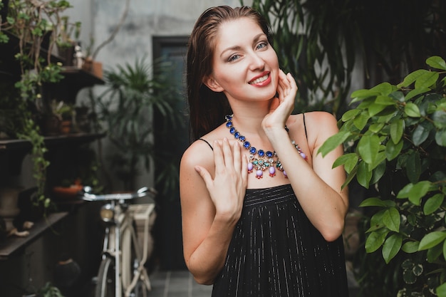 Portrait of young attractive woman in stylish black dress posing at tropical villa, sexy, elegant summer style, fashionable necklace accessories, smiling, jewelry, pure natural skin of face