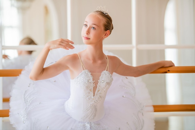 Portrait of a young ballerina in a beautiful white tutu near ballet barre in large beautiful white hall.