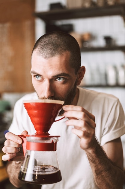 Портрет молодого бариста, стоящего у барной стойки и нюхающего кофе, задумчиво глядя в сторону в кафе