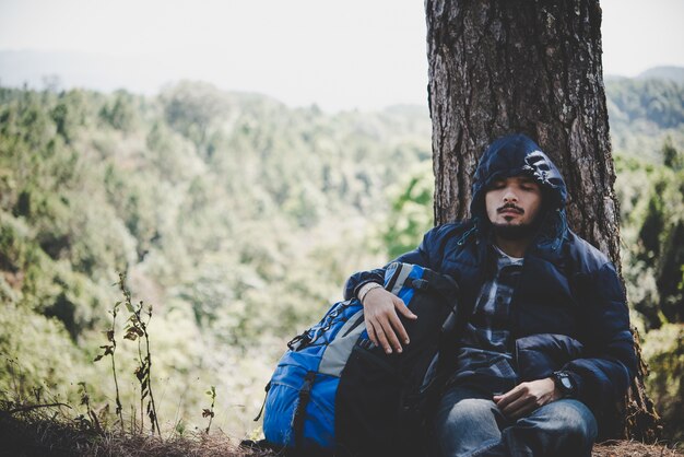 若い髭の男の肖像画は、離れて見えるバックパックと木々で単独で座って、自然と楽しむ。