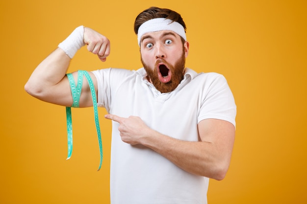 Portrait of a young bearded excited fitness man measuring biceps