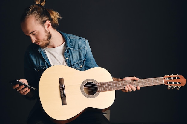 Foto gratuita ritratto di giovane musicista barbuto con chitarra sulle ginocchia utilizzando smartphone su sfondo nero. studio girato