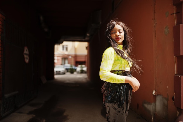 Портрет молодой красивой афро-американской фотомодели в зеленом топе и черных блестящих мокрых брюках на фоне арочного тоннеля
