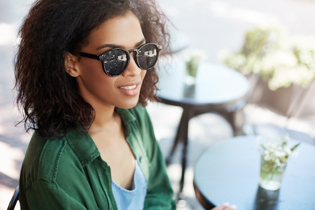 Ritratto di giovane bella donna africana in occhiali da sole sorridente che riposa rilassante nella caffetteria sulla terrazza.