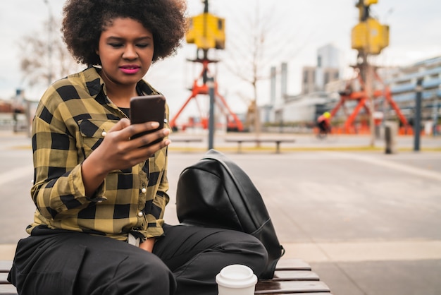 屋外のベンチに座っている間彼女の携帯電話を使用して若い美しいアフロアメリカンラテン女性の肖像画。コミュニケーションの概念。