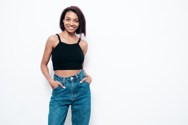 Portrait of young beautiful black woman Smiling model dressed in summer jeans and black top clothes Sexy carefree female posing near white wall in studio Tanned and cheerful