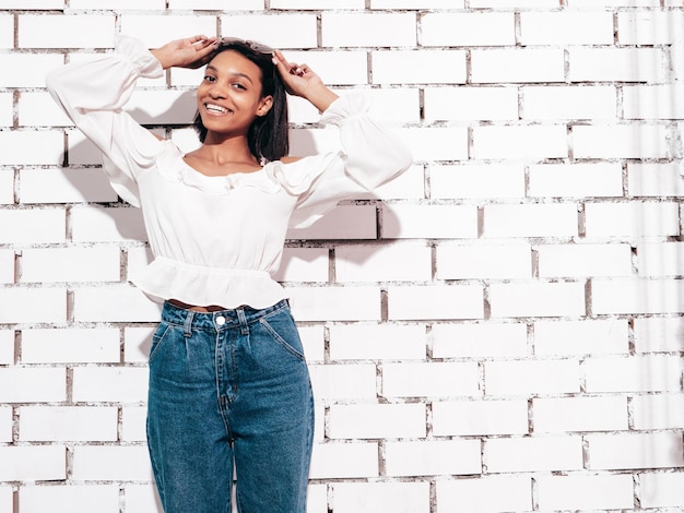 Portrait of young beautiful black woman Smiling model dressed in summer jeans clothes Sexy carefree female posing near white brick wall in studio Tanned and cheerful