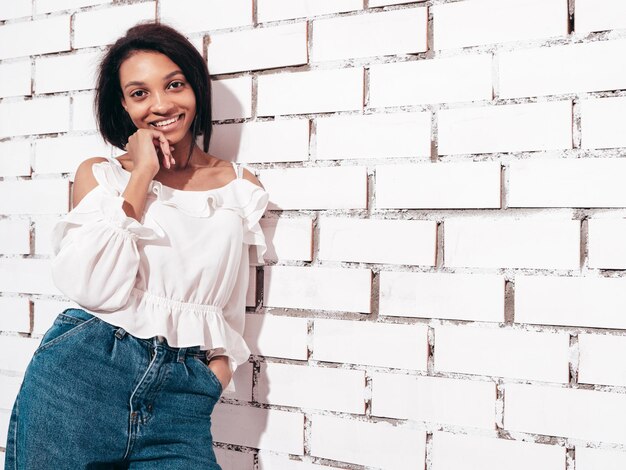 Portrait of young beautiful black woman Smiling model dressed in summer jeans clothes Sexy carefree female posing near white brick wall in studio Tanned and cheerful