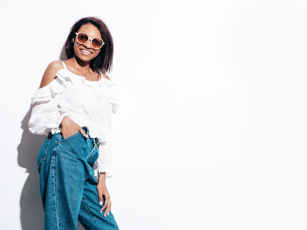 Portrait of young beautiful black woman Smiling model dressed in summer jeans clothes Sexy carefree female posing near white wall in studio Tanned and cheerful