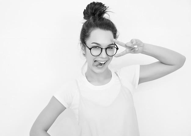 Portrait of young beautiful brunette woman girl model with nude makeup in colorful summer pink hipster clothes posing near wall. Showing her tongue and peace sign