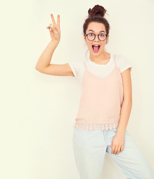 Portrait of young beautiful brunette woman girl model with nude makeup in colorful summer pink hipster clothes posing near wall. showing peace sign