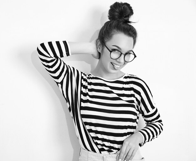 Portrait of young beautiful brunette woman girl model with nude makeup in glasses in summer hipster clothes posing near wall. showing her tongue