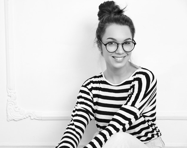 Portrait of young beautiful brunette woman girl model with nude makeup in glasses in summer hipster clothes posing near wall. Sitting on the floor