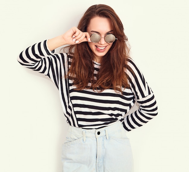 Portrait of young beautiful brunette woman girl model with nude makeup in summer hipster clothes and sunglasses posing near wall. showing her tongue