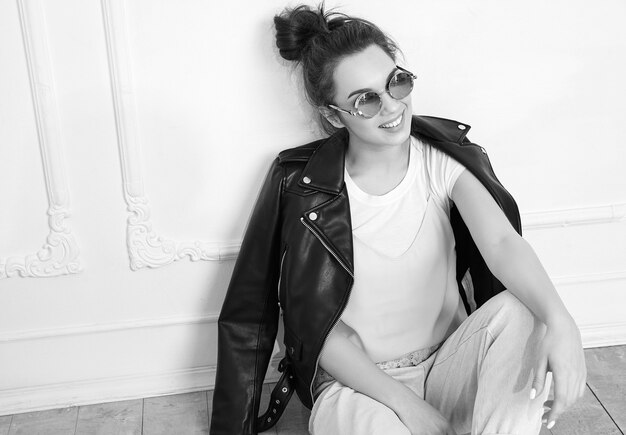 Portrait of young beautiful brunette woman girl model with nude makeup wearing summer hipster biker leather jacket clothes in sunglasses posing near wall. Sitting on the floor
