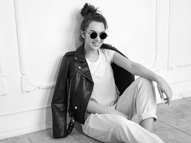 Portrait of young beautiful brunette woman girl model with nude makeup wearing summer hipster biker leather jacket clothes in sunglasses posing near wall. Sitting on the floor