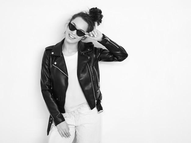 Portrait of young beautiful brunette woman girl model with nude makeup wearing summer hipster biker leather jacket clothes in sunglasses posing near wall. 