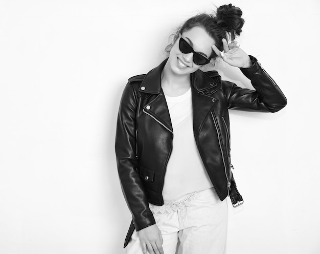 Portrait of young beautiful brunette woman girl model with nude makeup wearing summer hipster biker leather jacket clothes in sunglasses posing near wall. 
