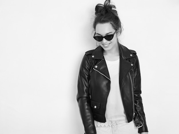 Portrait of young beautiful brunette woman girl model with nude makeup wearing summer hipster biker leather jacket clothes in sunglasses posing near wall. 