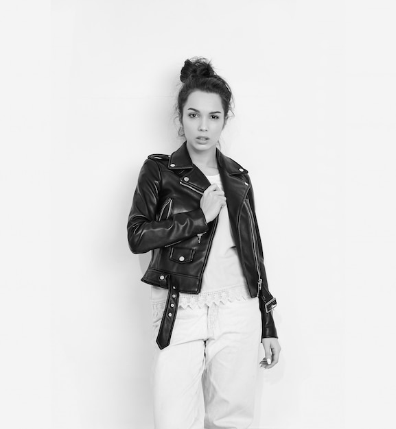 Portrait of young beautiful brunette woman girl model with nude makeup wearing summer hipster biker leather jacket posing near wall. 