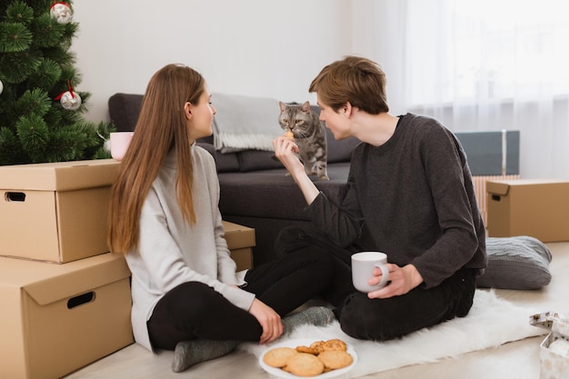 Portrait of young beautiful couple sitting on floor at home with cookies and looking at cat near