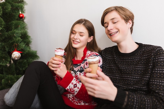 Ritratto di giovane bella coppia seduta sul divano a casa e felicemente trascorrendo del tempo insieme mentre mangiano il gelato con l'albero di natale vicino