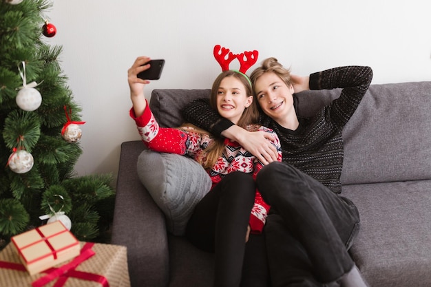 Portrait of young beautiful couple sitting on sofa at home and happily taking selfie together with Christmas tree near