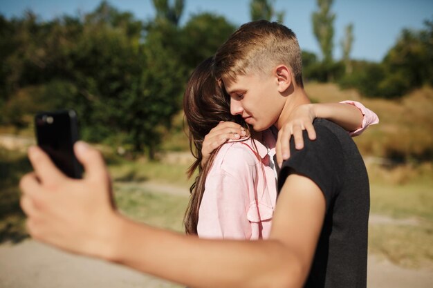 公園で自分撮りをしながら立って抱き合っている若い美しいカップルの肖像画。携帯電話の正面カメラで写真を撮るかわいいカップル