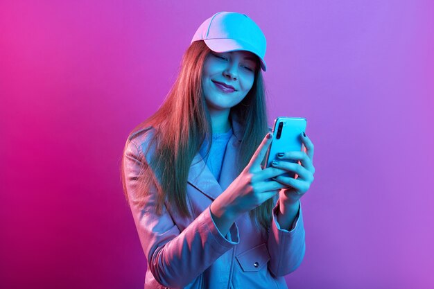 Портрет молодой красивой модной модели в кожаном пекаре и бейсболке, держа в руках смартфон