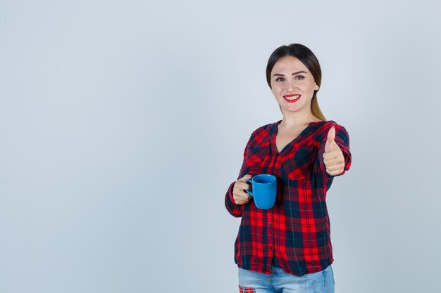 Portrait of young beautiful female holding cup while showing thumb up in casual shirt, jeans and looking blissful front view