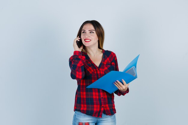 전화 통화를 하는 동안 폴더를 들고 있는 젊고 아름다운 여성의 초상화, 캐주얼 셔츠, 청바지를 입고 행복한 앞모습을 바라보는 모습