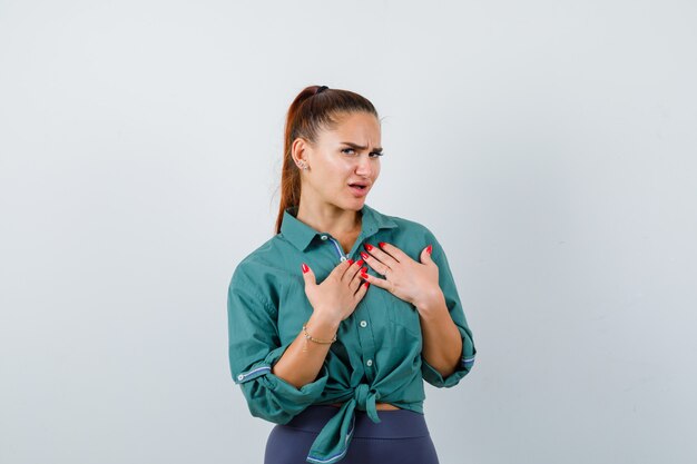 Portrait of young beautiful female keeping hands on chest in green shirt and looking indecisive front view