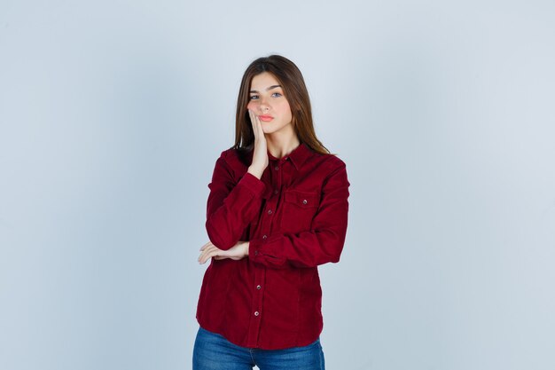 Portrait of young beautiful female leaning cheek on palm in shirt and looking pensive