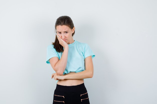 Portrait of young beautiful female leaning cheek on palm in t-shirt, pants and looking thoughtful front view