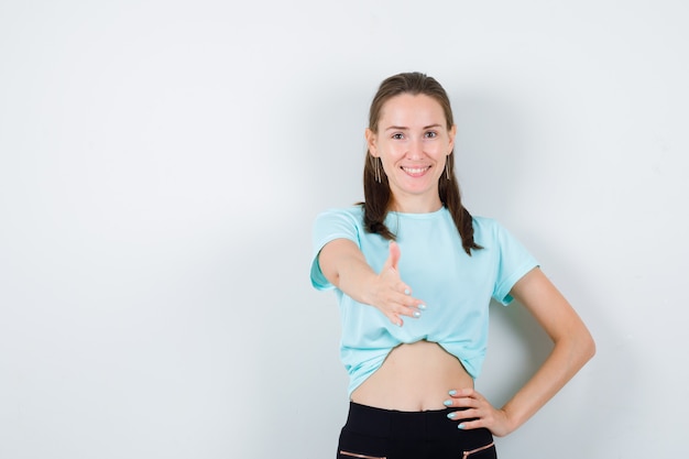 Ritratto di giovane bella donna che offre stretta di mano per salutare in maglietta, pantaloni e guardando allegra vista frontale