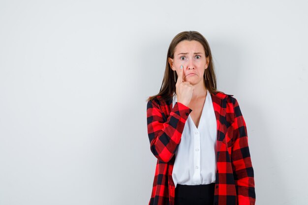 Portrait of young beautiful female pointing at her eye in casual outfit and looking sad front view