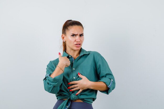 Portrait of young beautiful female pointing at herself while keeping hand on chest in green shirt and looking bewildered front view