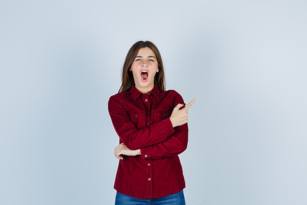 Portrait of young beautiful female pointing at upper right corner, yawning in shirt and looking sleepy