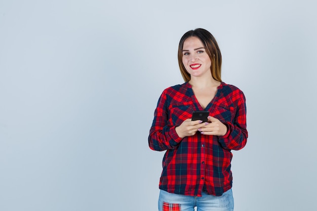 Portrait of young beautiful female posing while holding phone in casual shirt and looking blissful front view