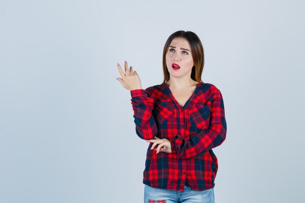 Portrait of young beautiful female raising hand in questioning manner, looking away in casual shirt and looking hesitant front view