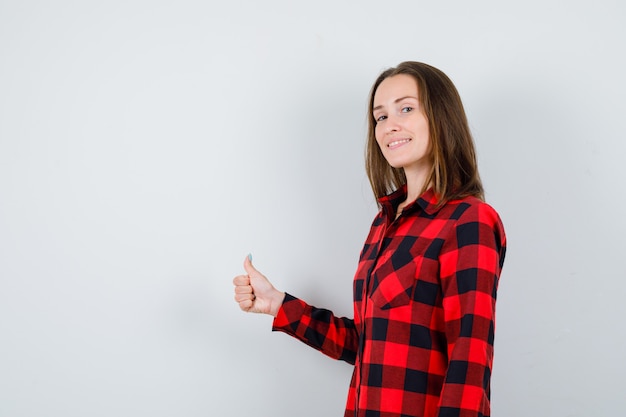 Portrait of young beautiful female showing thumb up in casual shirt and looking merry front view