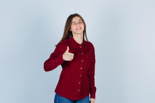 Portrait of young beautiful female showing thumb up, winking eye in shirt and looking merry