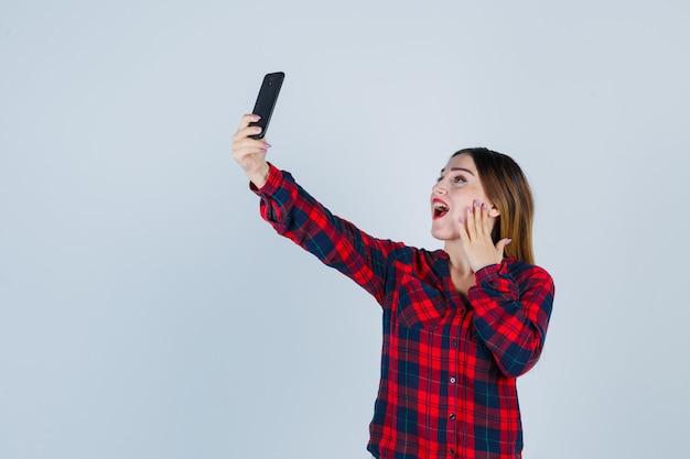 Ritratto di giovane bella donna che prende selfie, con la mano sulla guancia in camicia casual e sembra felice vista frontale